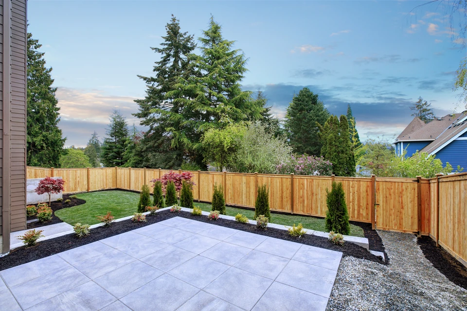 Jardin résidentiel moderne avec terrasse en dalles de béton, bordures paysagères en paillis noir, pelouse tondue et plantation d'arbustes conifères le long d'une clôture en bois