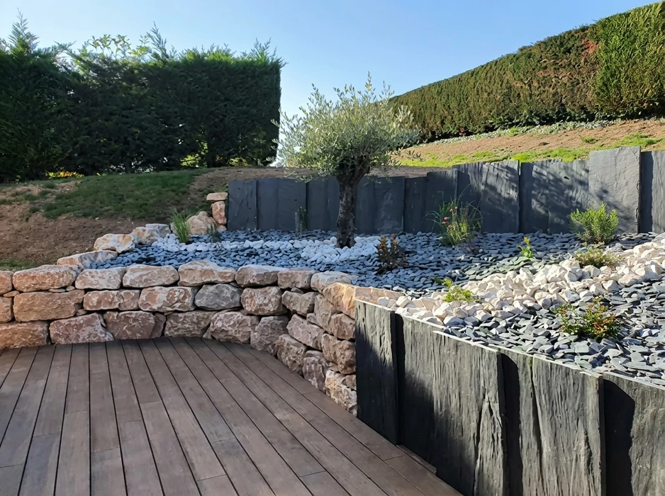 Aménagement paysager extérieur avec terrasse en bois composite, muret en pierre calcaire dorée à sec, bordures en ardoise noire verticale